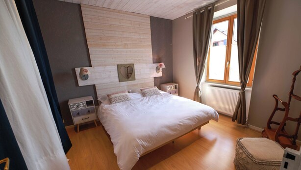 Une chambre à coucher avec une tête de lit en bois, un lit avec une literie blanche, une table de chevet avec une lampe, une fenêtre avec des rideaux et une chaise en bois.