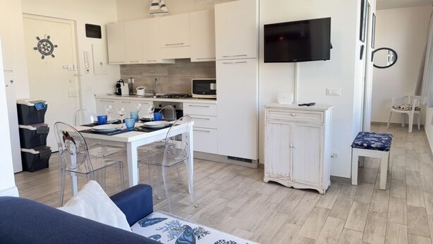 Ett kompakt kök med vita skåp, ett matbord med stolar, en tv som är monterad på väggen och en liten sittgrupp med en bänk.