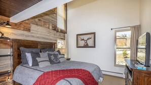 A bedroom with a wooden ceiling, a bed with grey bedding and a red throw, a mounted TV, and a window with a view of buildings. - Top Floor, End Unit Condo with Amazing Mountain Views (Silverthorne)