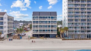 Un hôtel en bord de mer, avec plusieurs étages, des balcons et une plage de sable au premier plan.