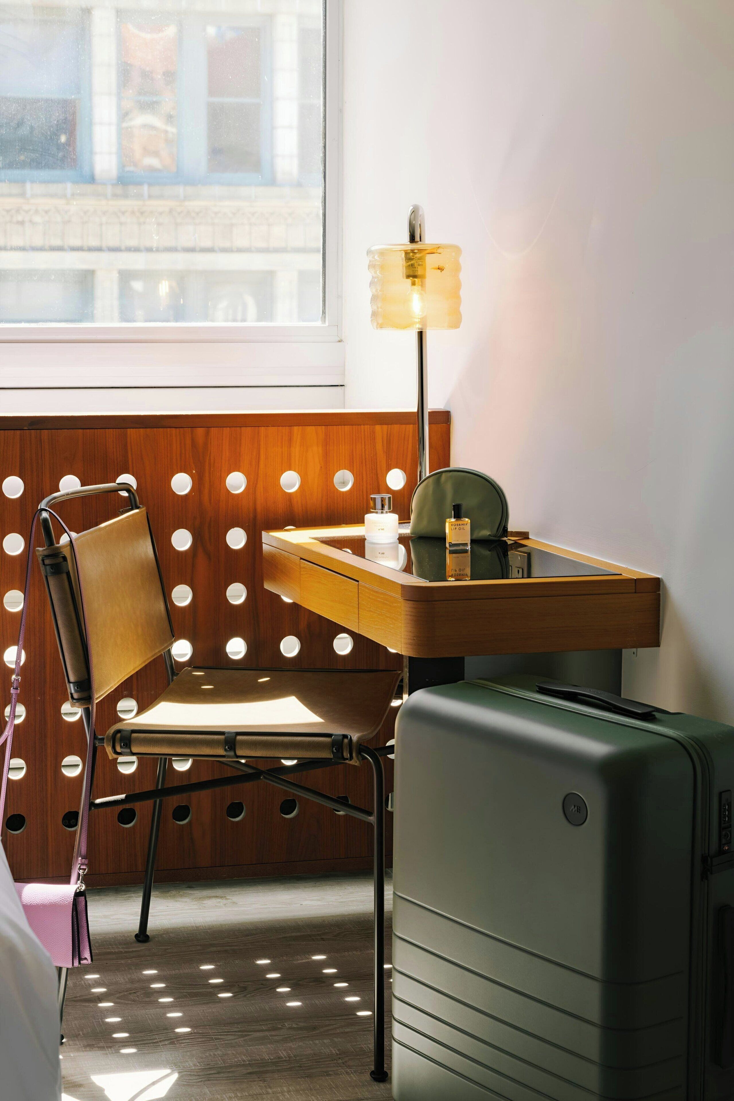 A hotel room with a wooden desk, a chair, a lamp, a suitcase, and a window with a view of buildings.