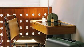 A hotel room with a wooden desk, a chair, a lamp, a suitcase, and a window with a view of buildings.