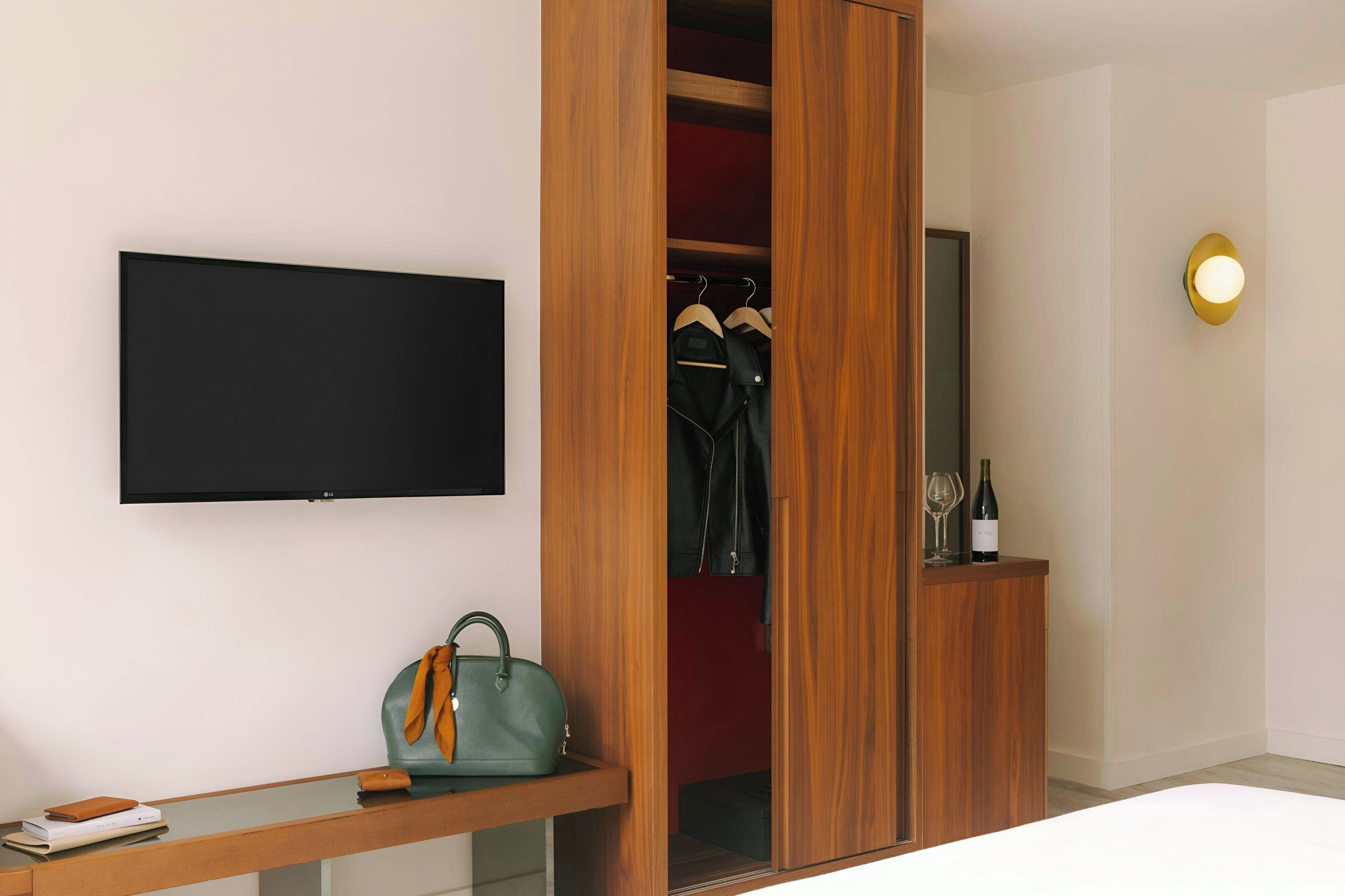 A modern room with a wooden wardrobe, a flat-screen TV, a desk with a handbag, and a wine bottle.