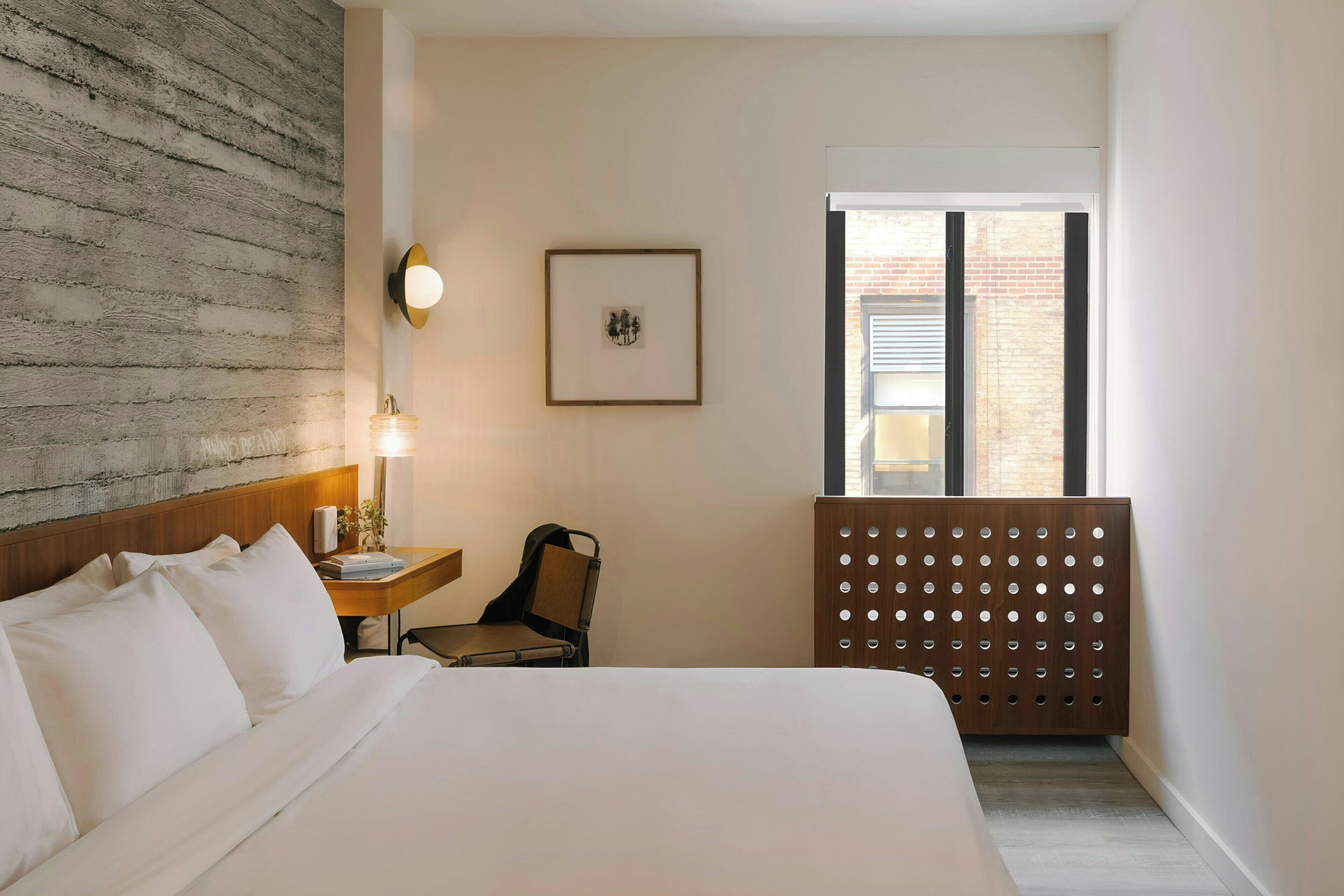 Habitación de hotel con una cama grande, cabecera de madera, escritorio con silla, lámpara de pared, cuadro enmarcado y ventana con persianas.