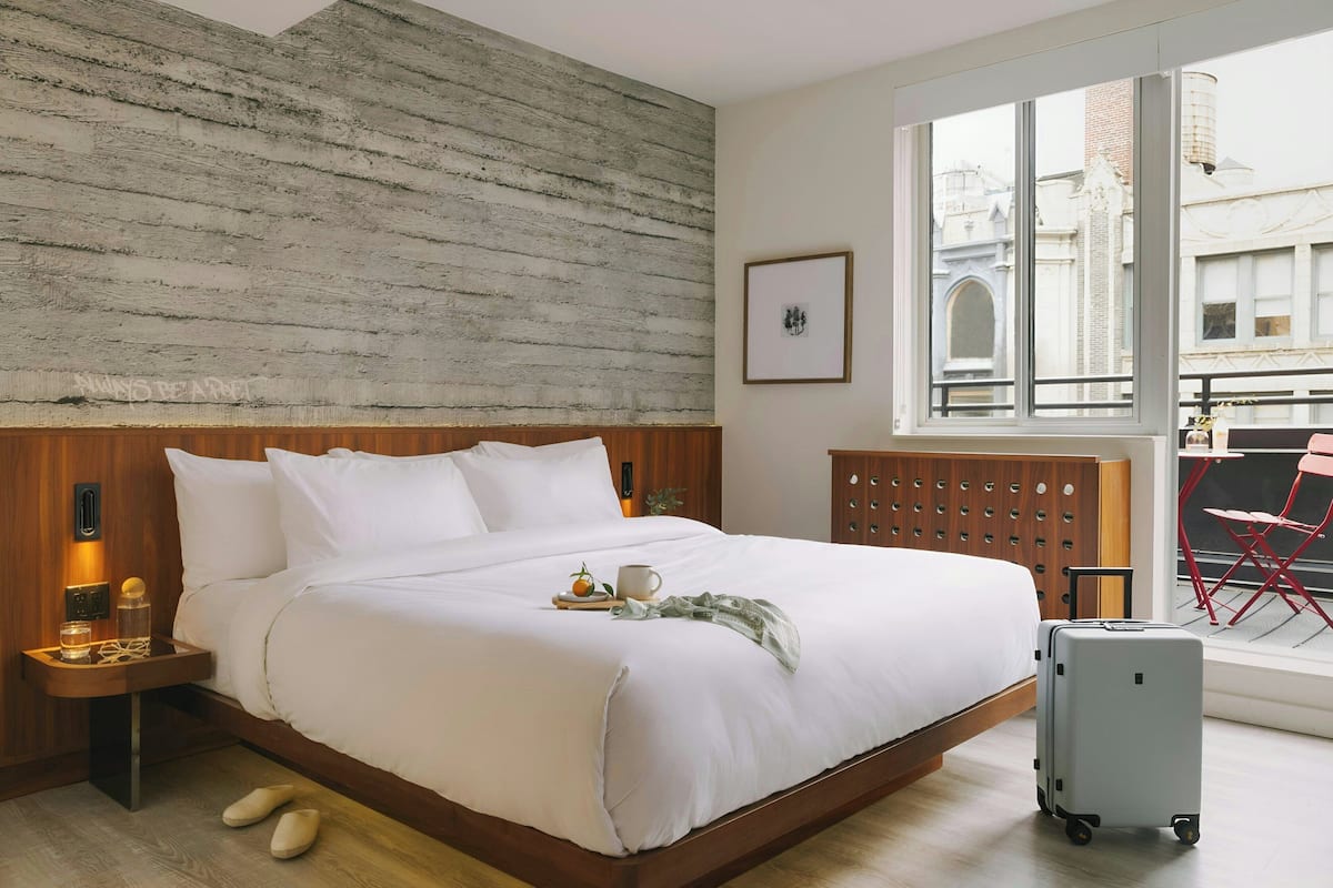 A neatly made bed with pillows and a tray, a suitcase, and a window with a view of buildings.