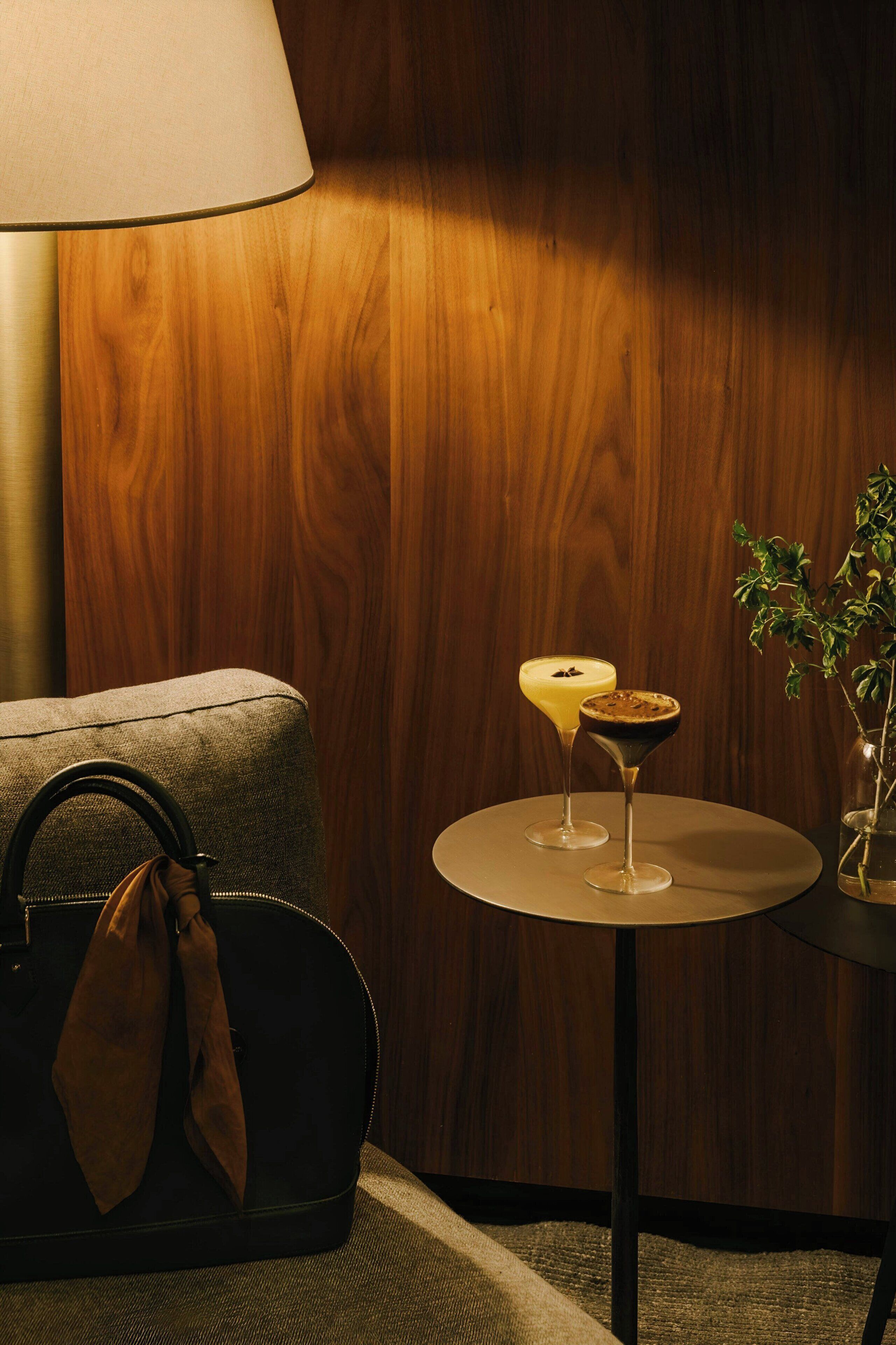 A wooden wall, a round table with two drinks, a lamp, and a handbag on a couch.