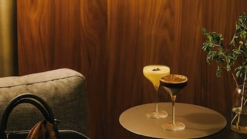 Una pared de madera, una mesa redonda con dos bebidas, una lámpara y un bolso sobre un sofá.