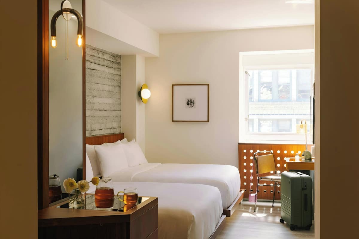 A hotel room with two beds, a desk with a lamp, a chair, a suitcase, and a window with a view.