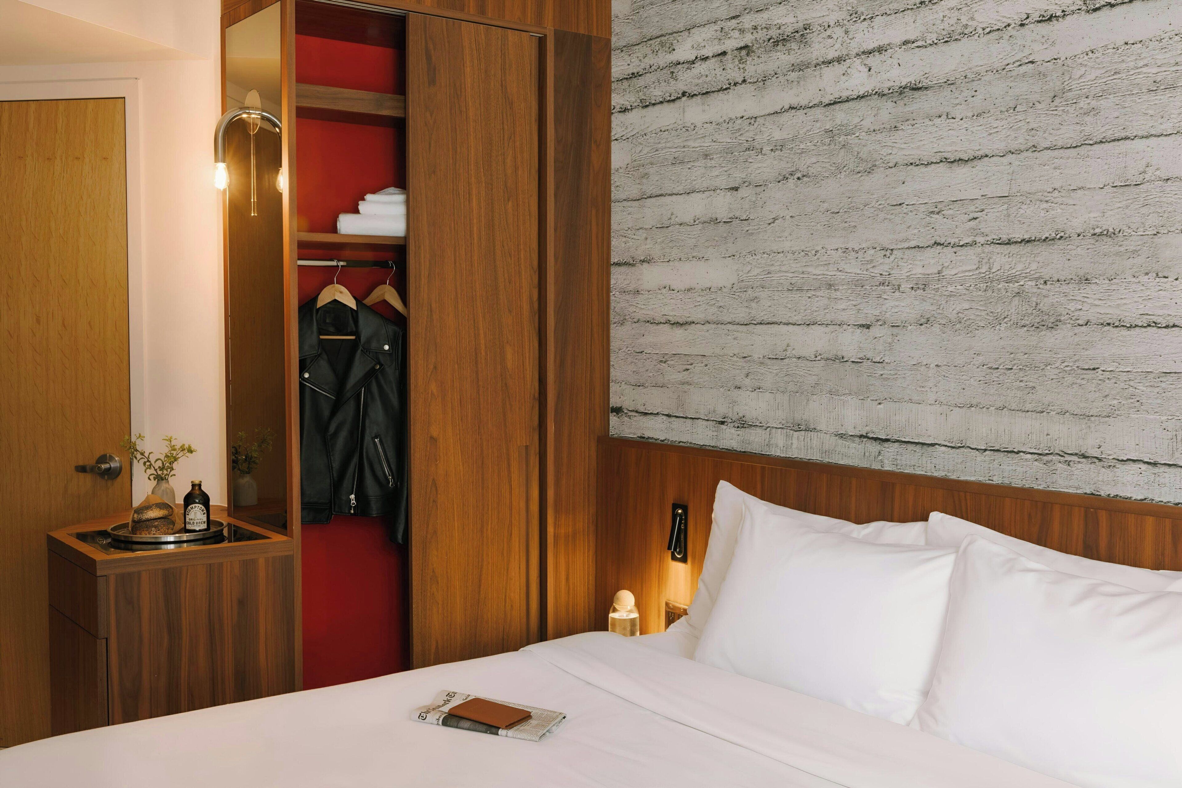 A hotel room with a wooden wardrobe, a bed with white linens, a red wall, and a textured wall behind the bed.