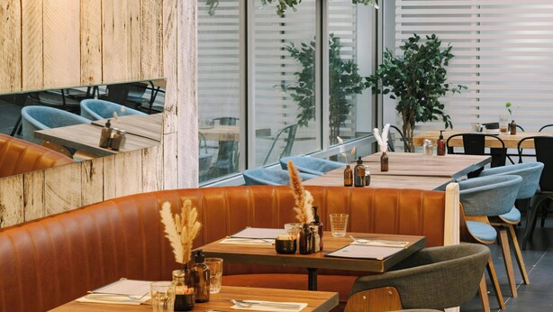 A restaurant with wooden tables and booths, large windows, and indoor plants. - Hotel Henri NY (New York)
