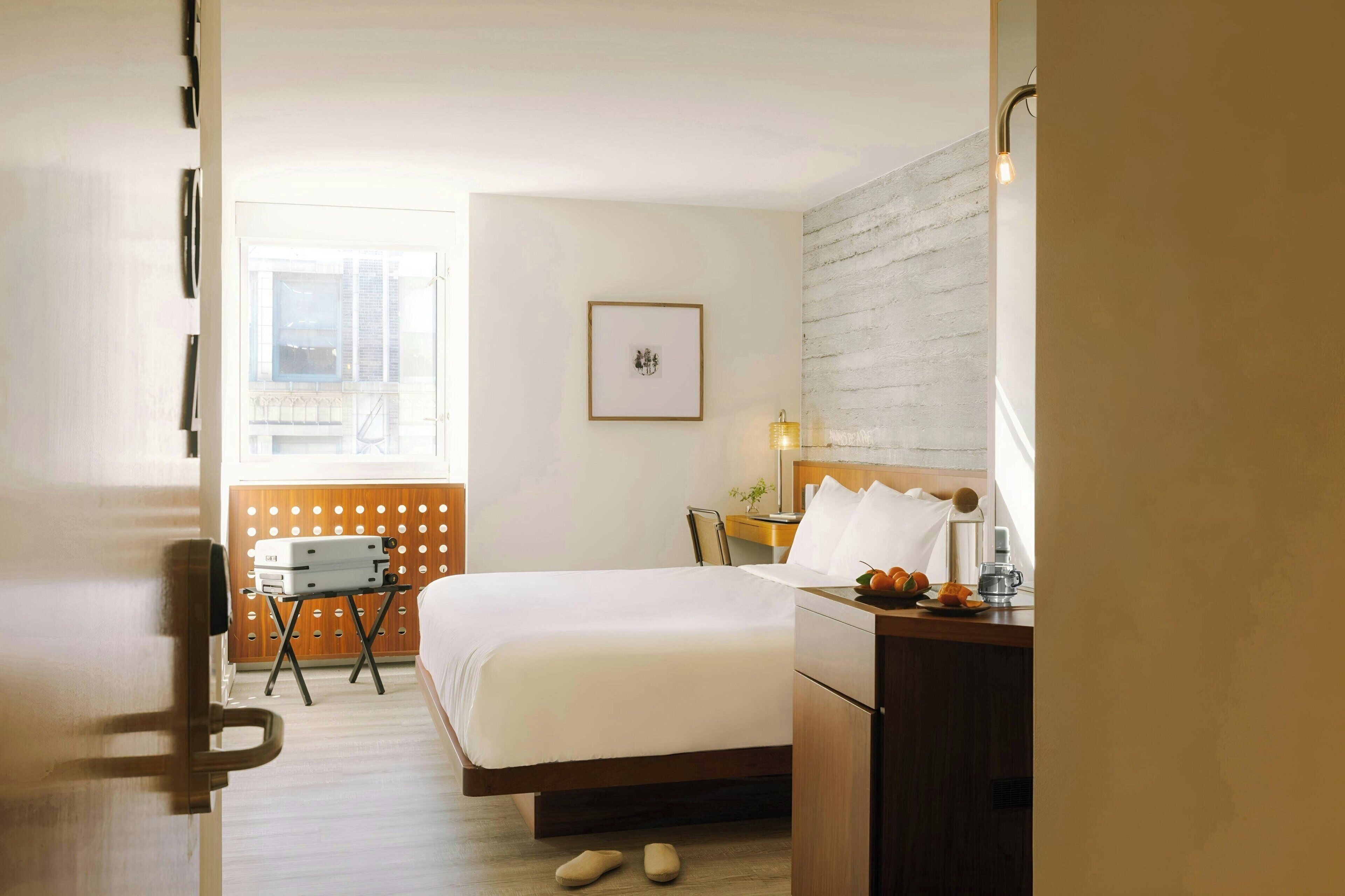 Quarto de hotel com cama, mesa de cabeceira, pequena escrivaninha, cadeira, janela e luminária na parede.