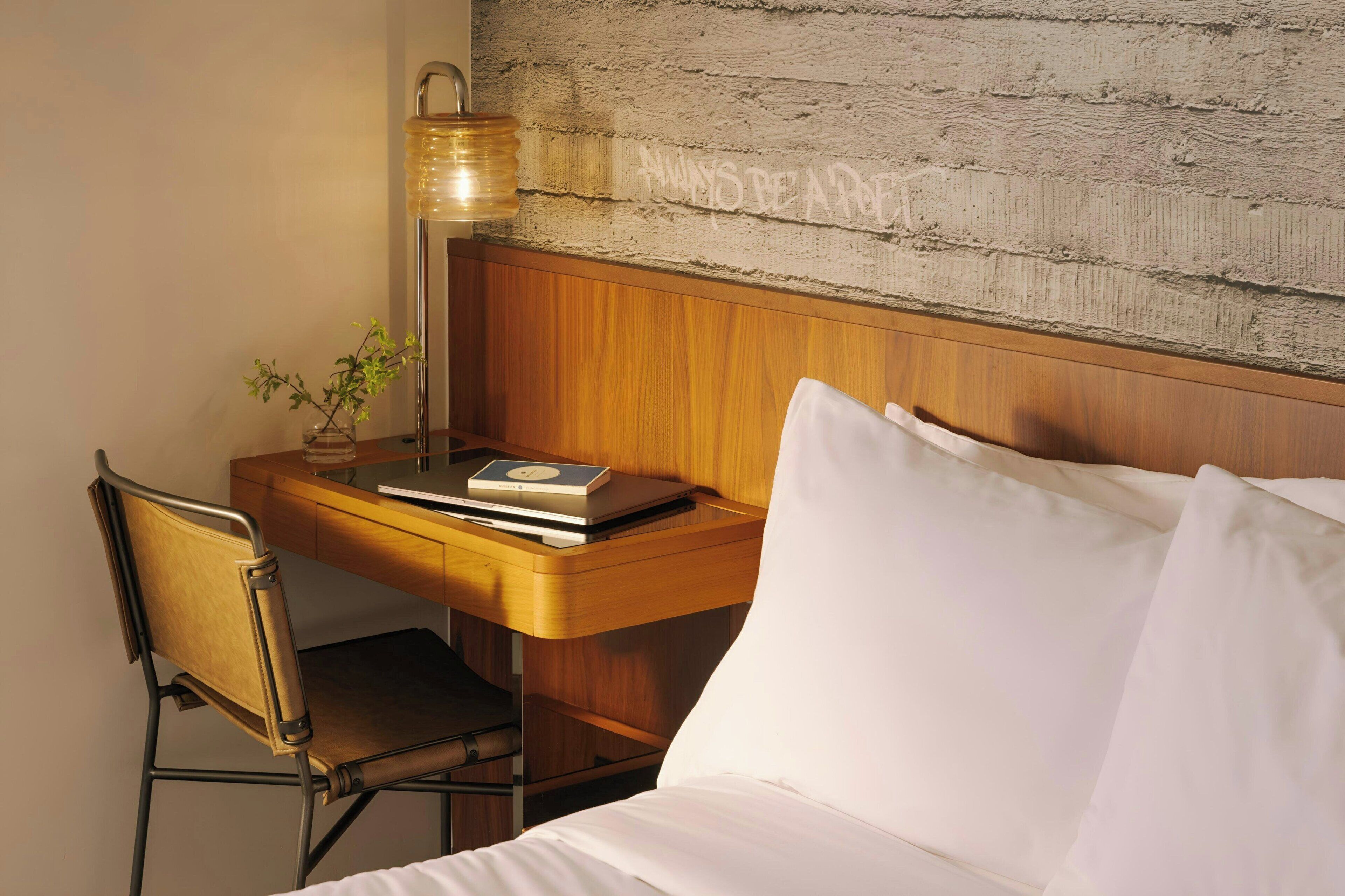 Ein ordentlich eingerichtetes Hotelzimmer mit einem Holztisch, einem Stuhl, einem Bett mit weißer Bettwäsche, einer Lampe und einem kleinen Topfpflanzen.