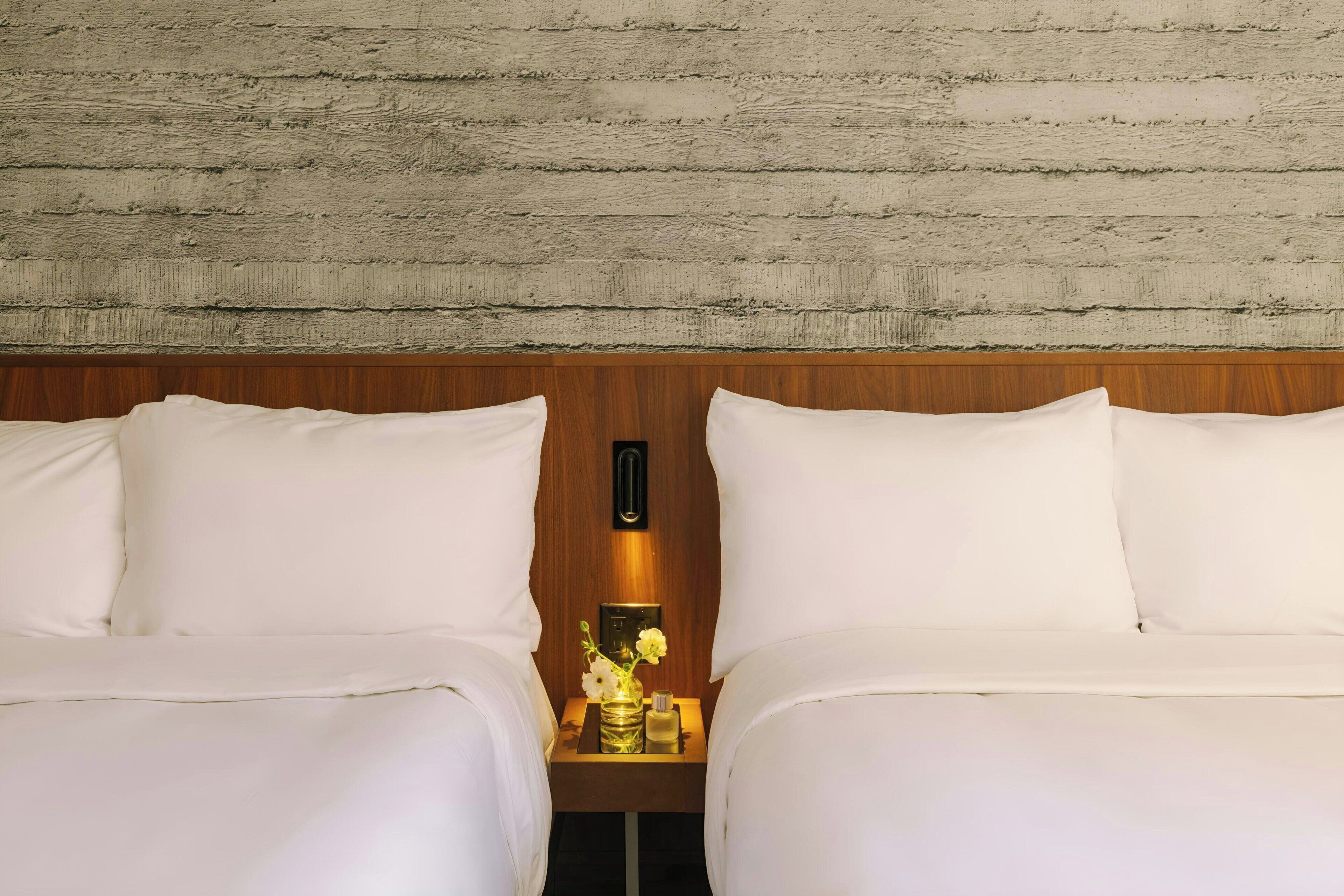 A hotel room with two beds, wooden headboard, a nightstand with a vase of flowers, and a wall with a textured pattern.