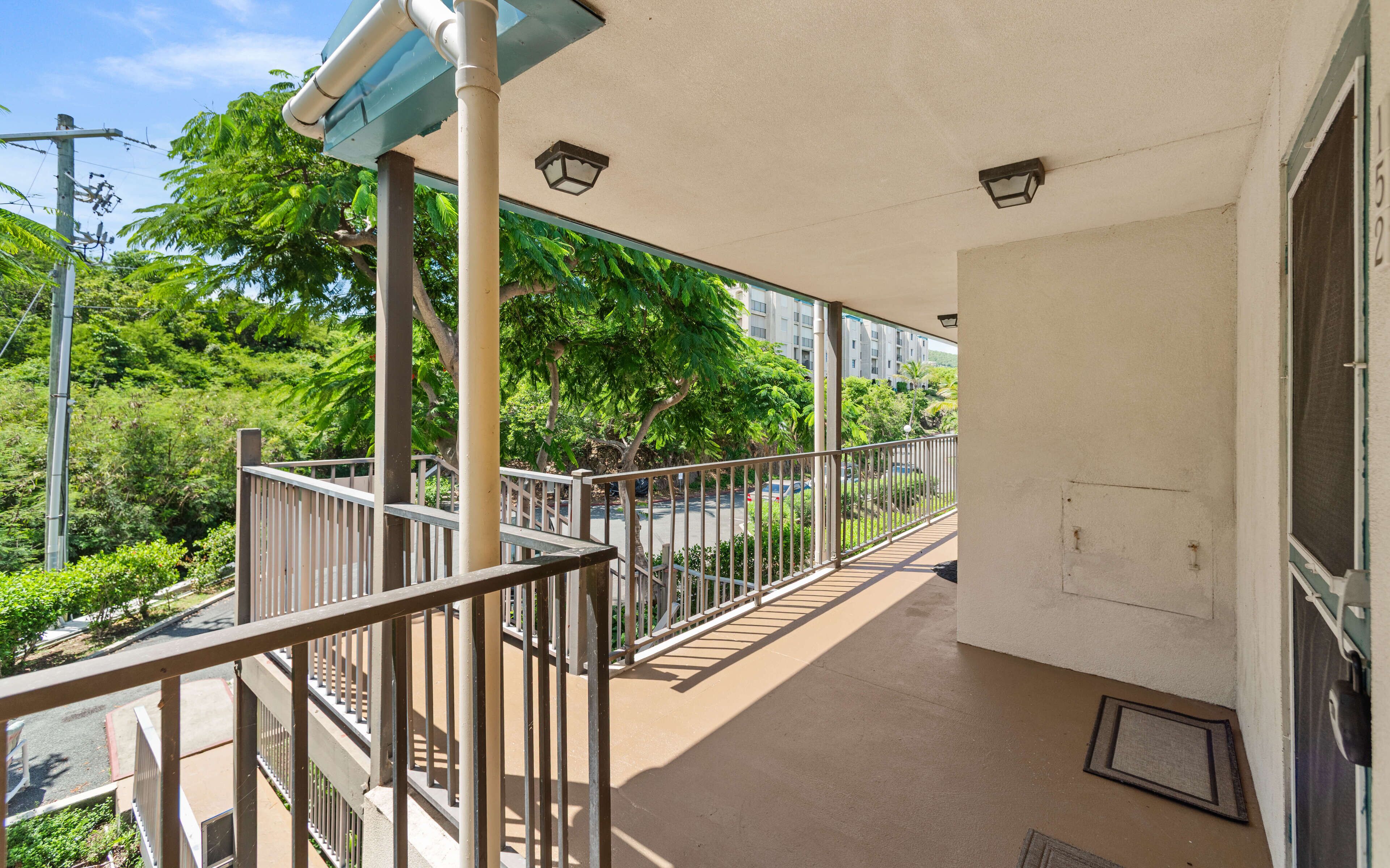 A balcony with a metal railing, a door, and a view of a parking area and greenery.