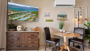 Een woonkamer met een televisie waarop een panoramisch uitzicht te zien is, een houten kast, twee leren fauteuils, een kleine tafel met een plant en een wandairconditioning.
