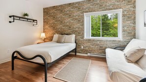 Un dormitorio con pared de piedra, ventana con vistas a árboles, cama, silla y estante pequeño con una planta.