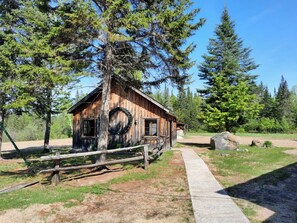Une cabane en bois rustique, dotée d’une fenêtre circulaire, entourée d’arbres et bordée d’un chemin de gravier.