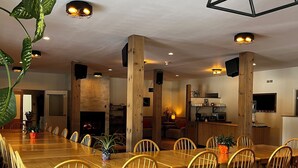 Un comedor amplio con mesas y sillas de madera, rodeado de plantas en macetas y con vigas de madera a la vista e iluminación empotrada.