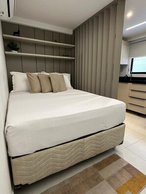 A modern bedroom with a bed, built-in shelving, and a kitchen area. - Aurea 210 (João Pessoa)