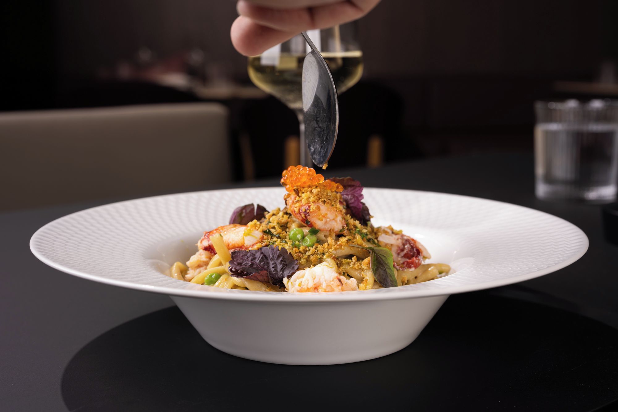 A white plate with a seafood dish, a spoon, and a glass of white wine on a table.