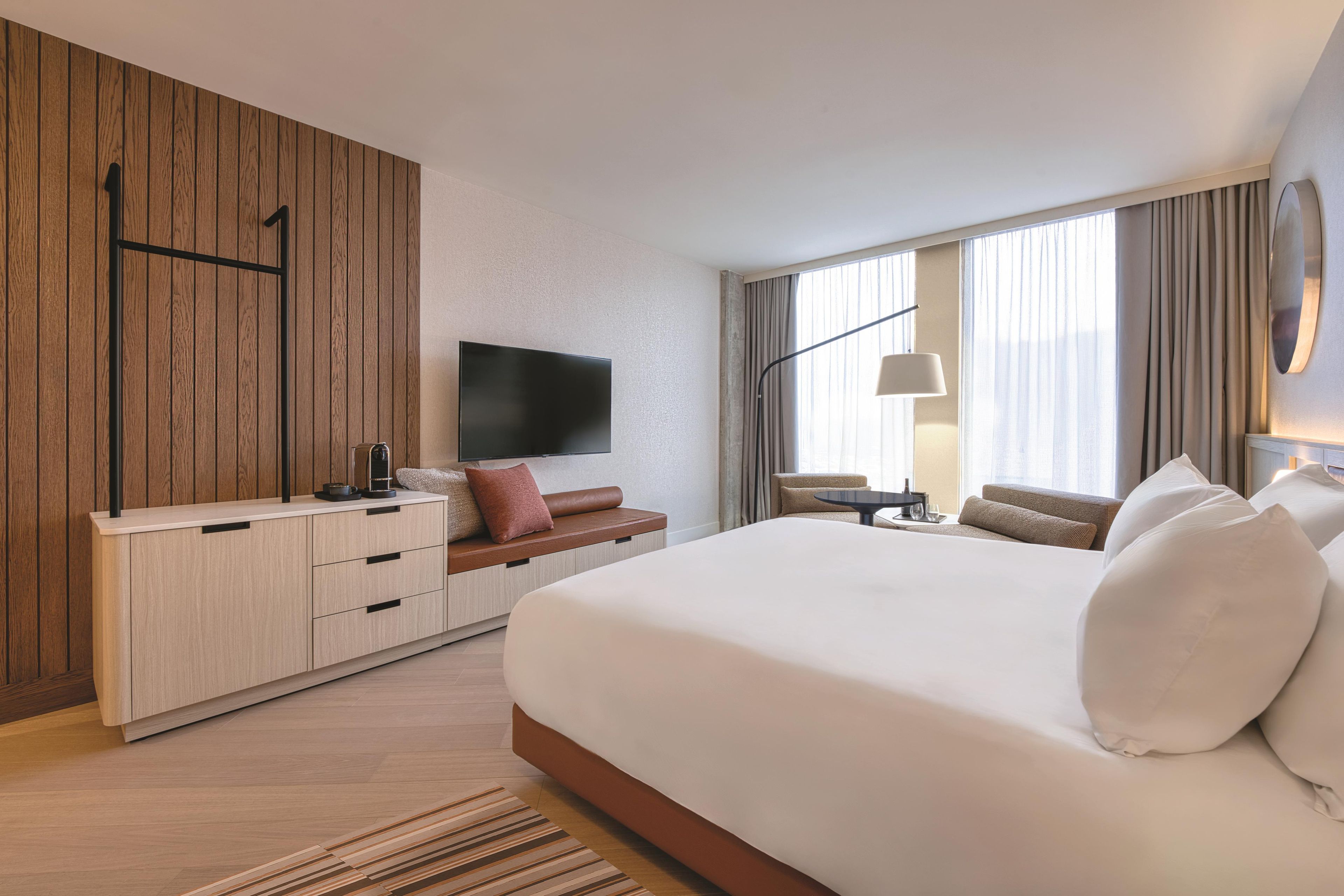 A hotel room with a large bed, a TV mounted on the wall, a desk with a lamp, and a seating area with a sofa and armchairs.