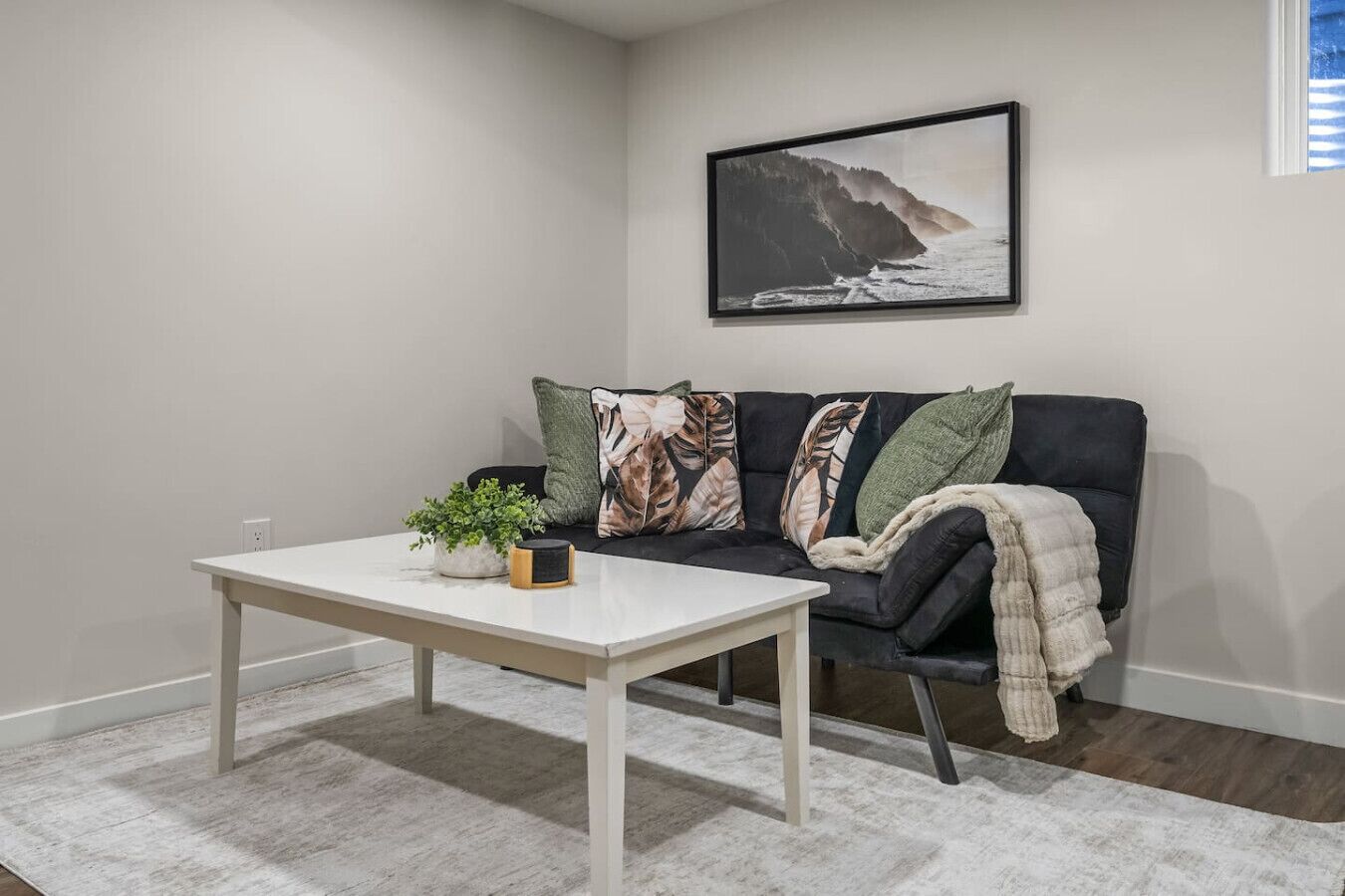A corner room with a white table, a black sofa with patterned cushions, a framed picture on the wall, and a small plant on the table.