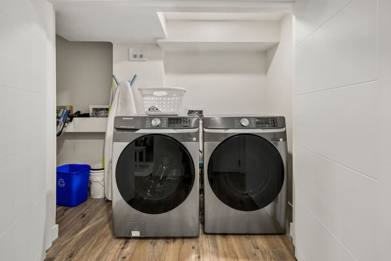 Una sala de lavandería con dos lavadoras de carga frontal, un contenedor azul para reciclaje y un estante blanco con varios objetos.