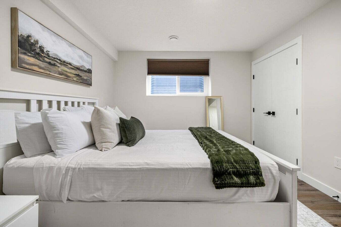 Ein Schlafzimmer mit einem weißen Bett, einem Fenster mit Jalousien, einem Spiegel und einer Tür.