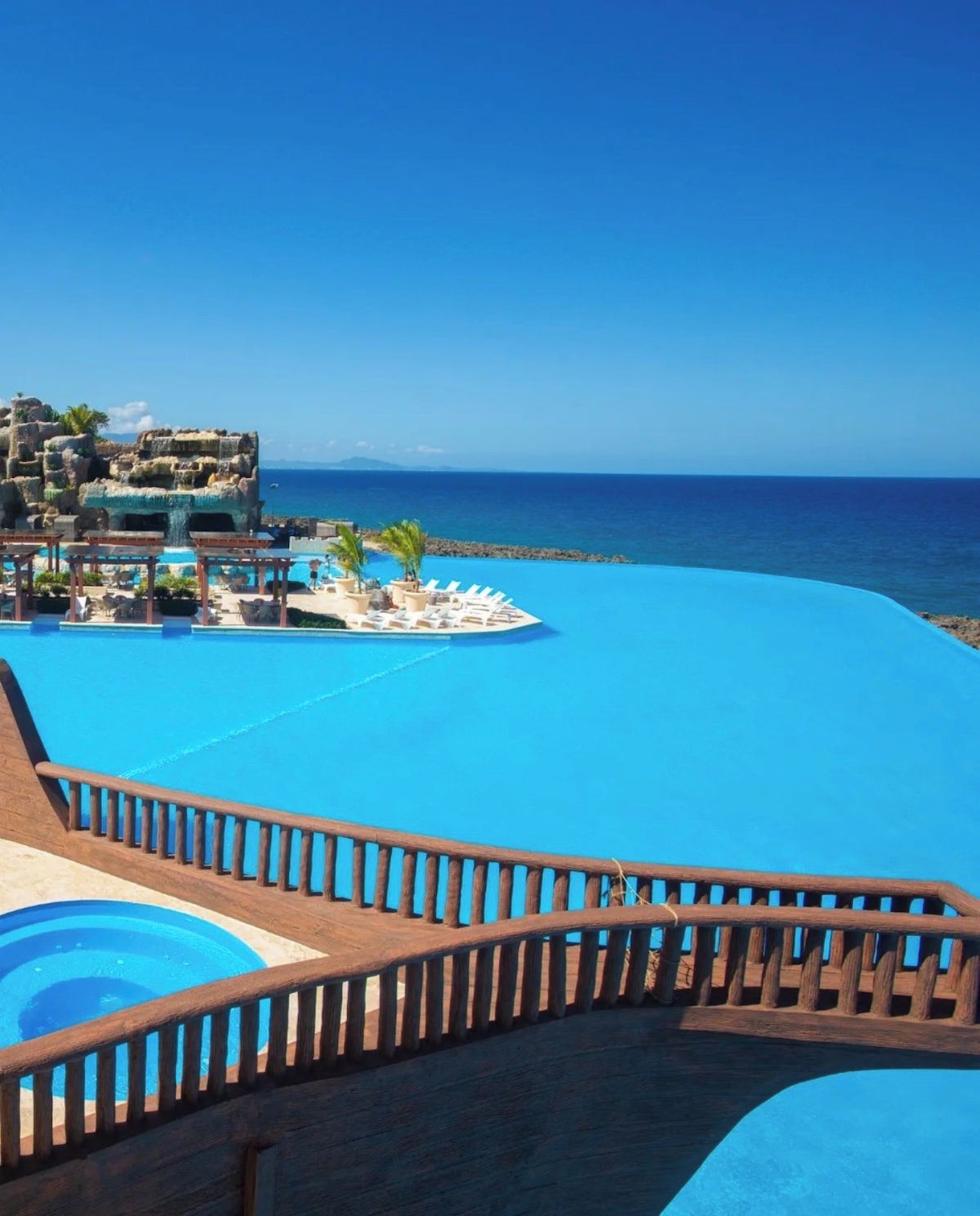 Une piscine d’un complexe hôtelier, dotée d’une terrasse en bois et de chaises longues, avec vue sur une mer d’un bleu limpide.