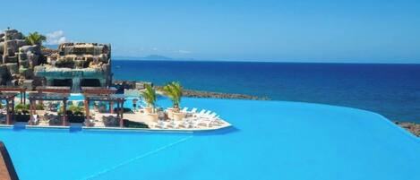 Une piscine d’un complexe hôtelier, dotée d’une terrasse en bois et de chaises longues, avec vue sur une mer d’un bleu limpide.