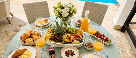 A round table set for breakfast with various dishes and drinks, overlooking a swimming pool and palm trees.
