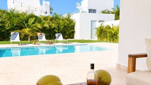 Una mesa de madera con una botella, vasos, cocos y un libro, con vista a una piscina y palmeras.