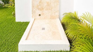 Una ducha exterior moderna con fachada de piedra, rodeada por un césped cuidado y plantas tropicales.