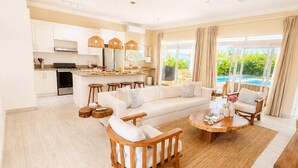 Amplia sala de estar con sofá, sillas y una mesa de centro. Se ve una cocina con armarios blancos y un comedor con una mesa y sillas.