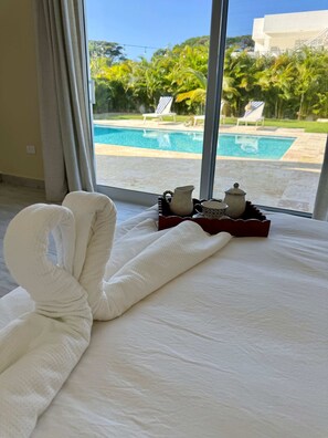 A bed with white towels, a tray with a pitcher and cups, and a view of a pool and palm trees. - Seahaven Villa in Oceanfront Comm Sosua Ocean Vill (Sosúa)