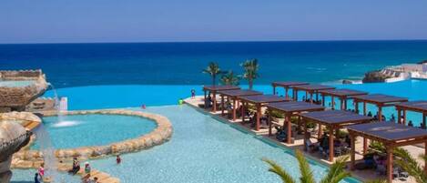 A resort pool area with clear blue water, a circular water feature, and lounge areas with umbrellas.
