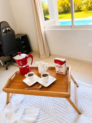 Uma bandeja de madeira com duas xícaras de café, uma cafeteira vermelha, um jarro branco e um saco de leite sobre um edredom branco acolchoado.