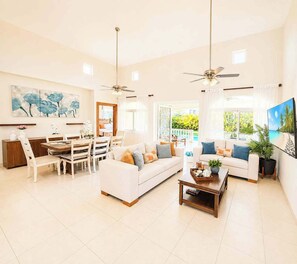 A spacious living area with a dining table, sofas, and a coffee table. There are ceiling fans, a large painting, and a view of the outdoors. - Sleeps up to 14 | 4 King Suites & Private Pool (Sosúa)