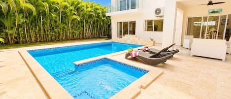 A modern pool area with a clear blue pool, lounge chairs, and a white building with large windows.
