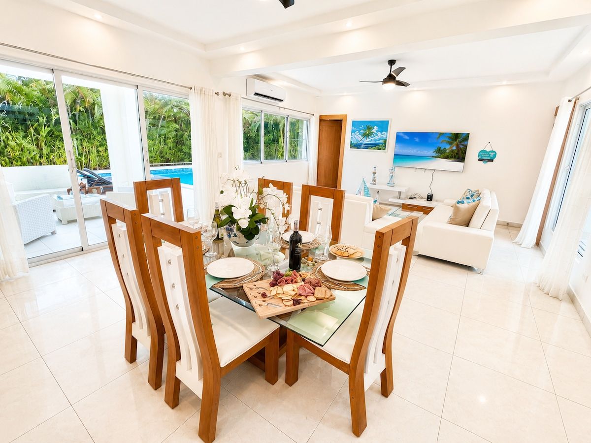 Un coin repas avec une table en verre entourée de chaises en bois, un canapé blanc et une vue sur une piscine et la verdure environnante.