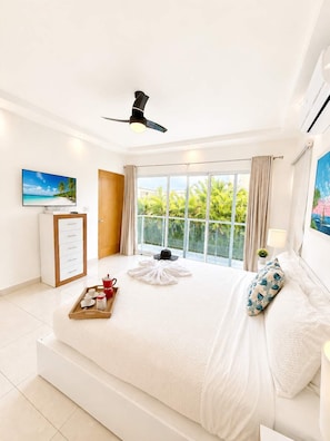 A hotel room with a large bed, a ceiling fan, a TV, a window with curtains, and a view of greenery outside. - 5bd Corner Lot Villa|Private Pool, Sleeps up to 14 (Sosúa)