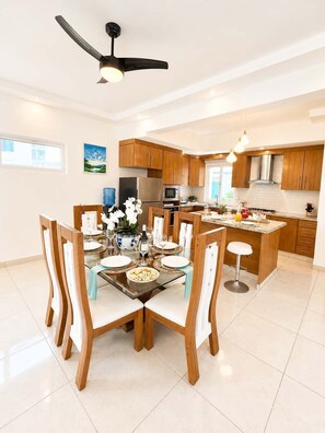 A modern kitchen with wooden cabinets, a central island, and a dining area with a glass table and chairs. - 5bd Corner Lot Villa|Private Pool, Sleeps up to 14 (Sosúa)