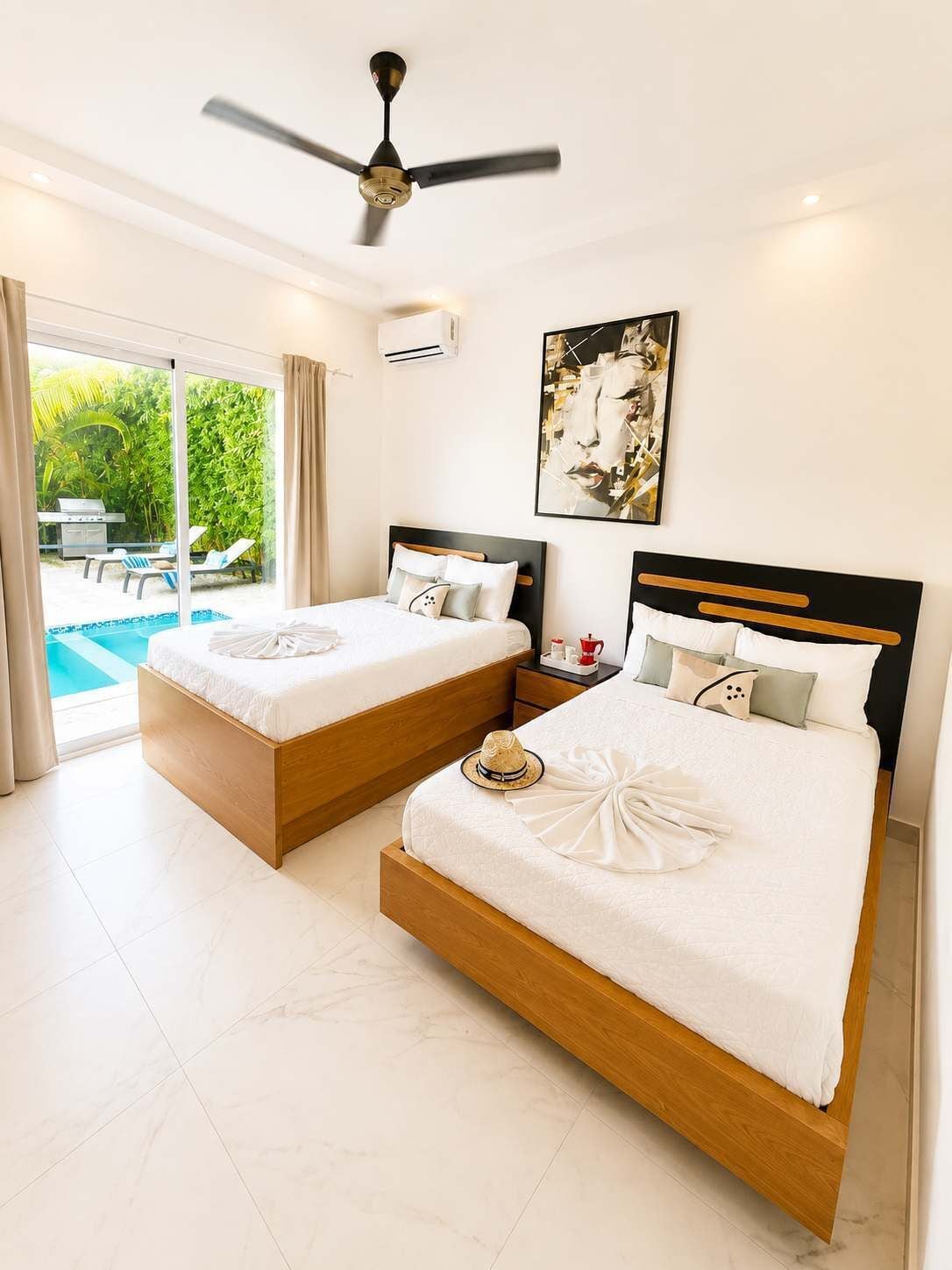 Habitación de hotel con dos camas, ventilador de techo, un cuadro y vistas a una piscina y zonas verdes.