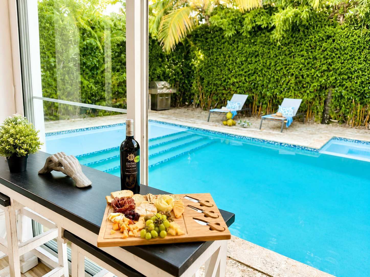 Blick auf ein Schwimmbecken mit einem Holztisch, auf dem sich eine Weinflasche, Käse und Trauben befinden.