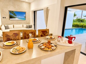 A breakfast setup with various pastries, a coffee pot, and a glass of orange juice on a table with a view of a swimming pool. - Bali Villa | Sunken Lounge & 3 King Beds in SOV (Sosúa)