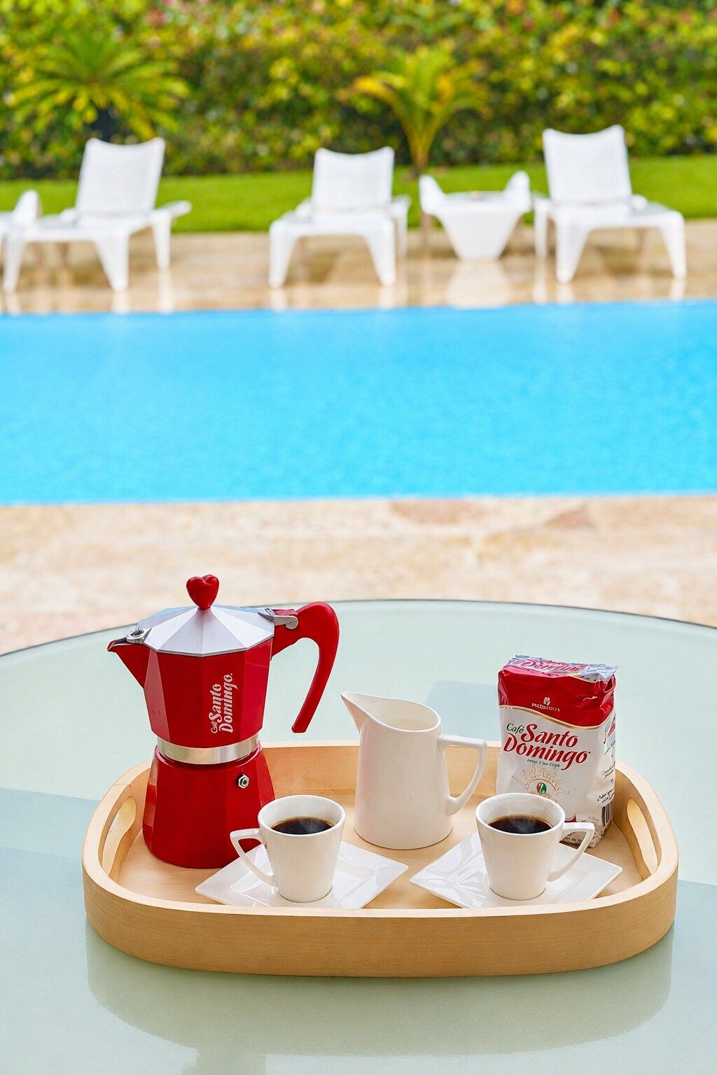 A table with a coffee setup including a red coffee pot, a white pitcher, and a package of coffee, with a pool and white chairs in the background.