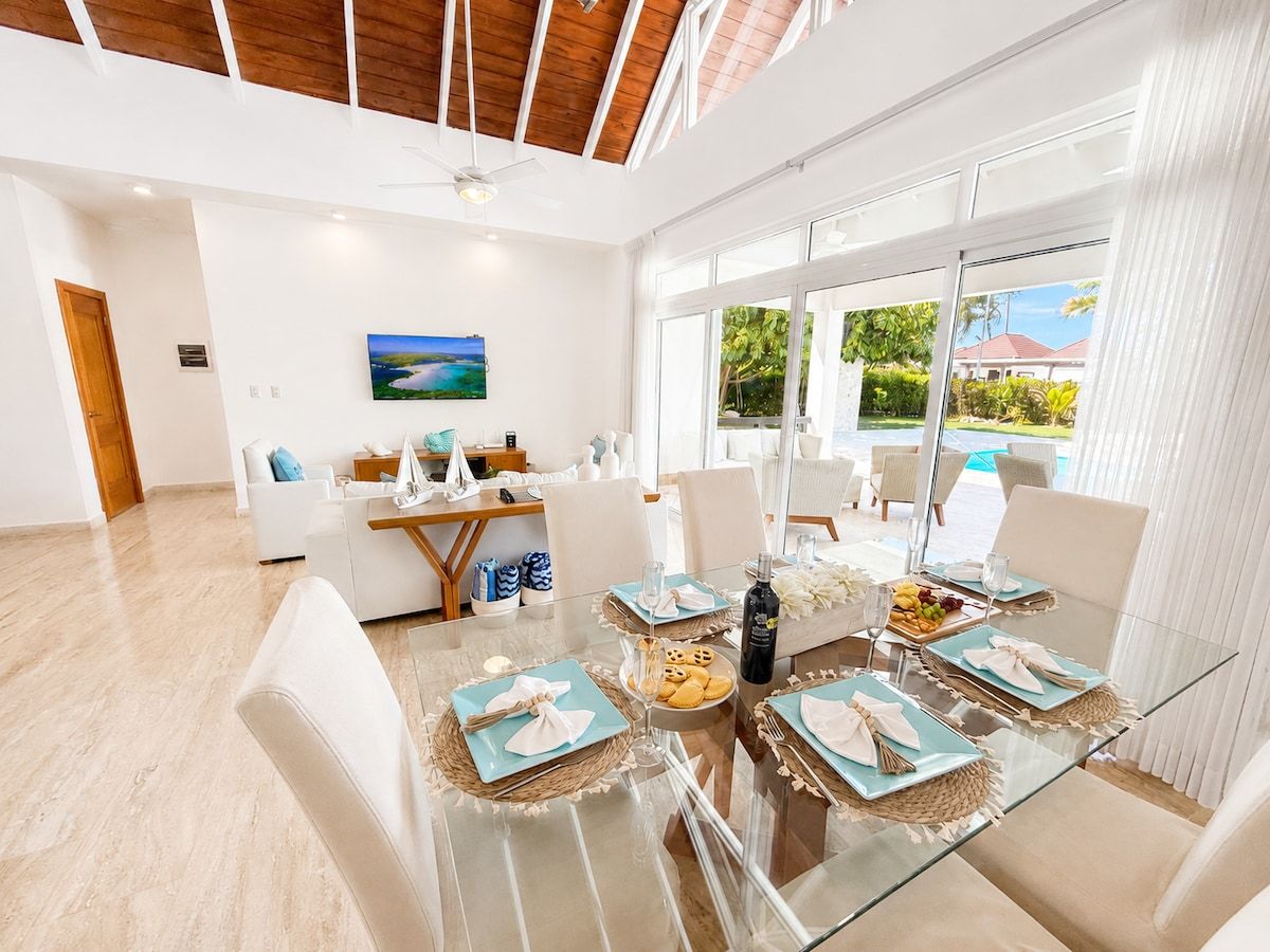 Uma área de jantar moderna com mesa de vidro posta para seis pessoas, cadeiras brancas e vista para o exterior.