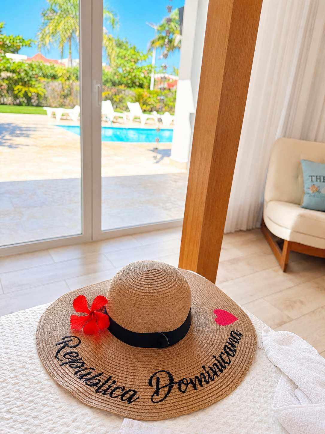 A straw hat with embroidered text on a textured surface, a white towel, and a view of a pool and greenery through a glass door.