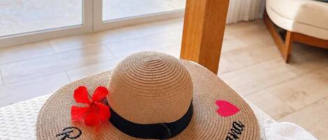 A straw hat with embroidered text on a textured surface, a white towel, and a view of a pool and greenery through a glass door.