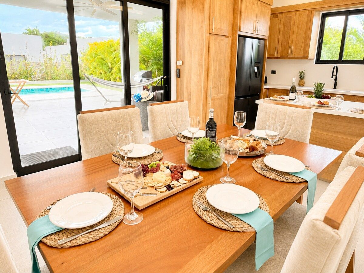 A dining area with a wooden table set for a meal, surrounded by chairs, and a view of a pool and greenery outside.